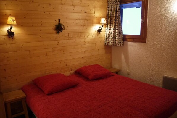 Une chambre avec une tête de lit en bois, deux lampes de chevet, une fenêtre avec des rideaux à motifs et une couverture de lit rouge.
