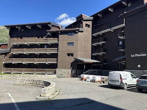 A modern building with multiple floors and balconies, a parking area with several cars, and a stone structure in the foreground.