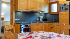 A compact kitchen with wooden cabinets, a stove, a coffee maker, and a table with a red and white patterned tablecloth.
