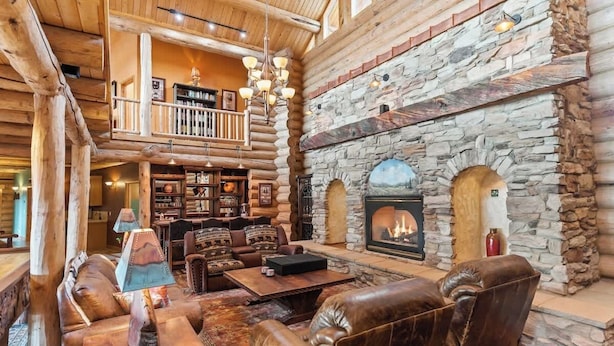 A log cabin interior with a stone fireplace, leather furniture, and a balcony overlooking the living area.