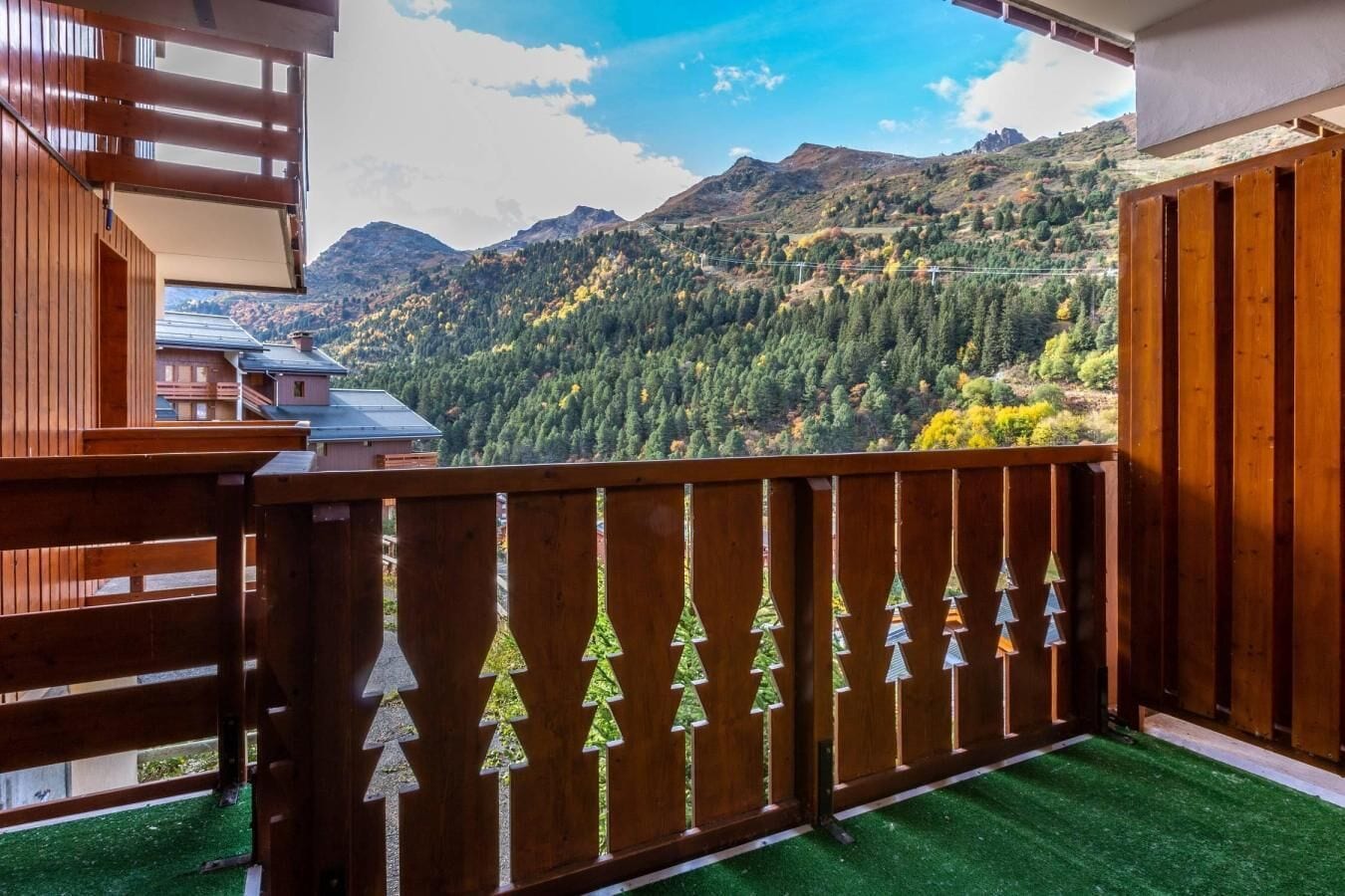 A balcony with a view of a mountainous landscape, a wooden fence with tree-shaped cutouts, and artificial green grass.