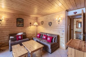 A cozy wooden cabin interior with a sofa set, two wooden tables, and wall-mounted lamps.