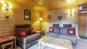 A cozy cabin interior with wooden walls, two sofas with heart-patterned cushions, a wooden coffee table, and wall-mounted lamps.