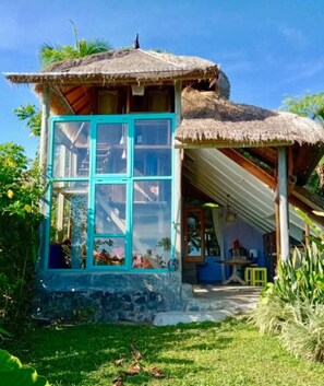 A rustic house with a thatched roof, a stone foundation, and a large window with blue trim. - Villa Pondok Apsari (Sukasada)