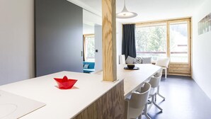 Une cuisine moderne avec un plan de travail blanc, une colonne en bois et un objet décoratif rouge. On y trouve une fenêtre donnant sur des arbres et un balcon avec une chaise bleue.