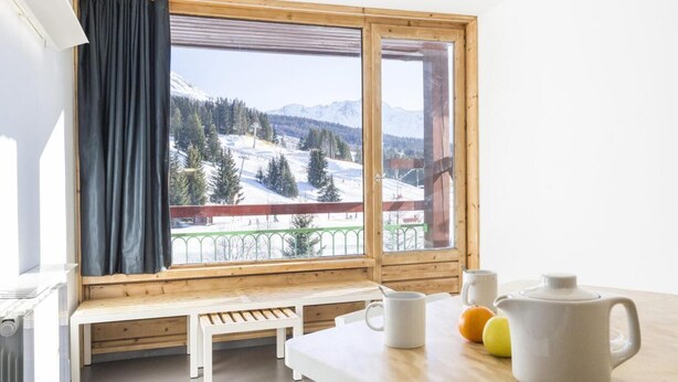 Une pièce avec un banc et une table en bois, une vue sur un paysage montagneux enneigé et une fenêtre avec un rideau.