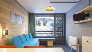 Una habitación moderna con pared de madera, sofá azul, banca, mesa, silla, televisor y ventana con vista nevada.