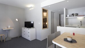 A compact hotel room with a TV, a small desk, a dining table, and a kitchenette.