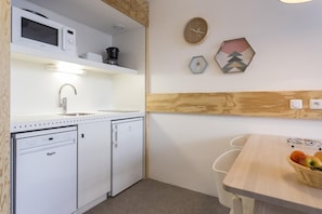 Une cuisine compacte équipée d’un four à micro-ondes, d’une machine à café et d’un lave-vaisselle. Un panneau mural en bois sépare la cuisine d’un coin repas comprenant une table et des chaises.