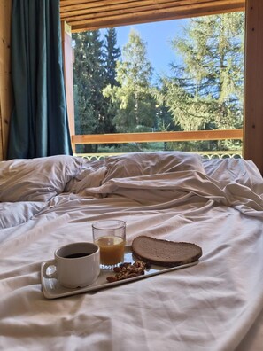 Ein Bett mit Tablett, auf dem eine Tasse, ein Glas Saft und eine Scheibe Brot stehen, mit Blick auf Bäume im Freien.