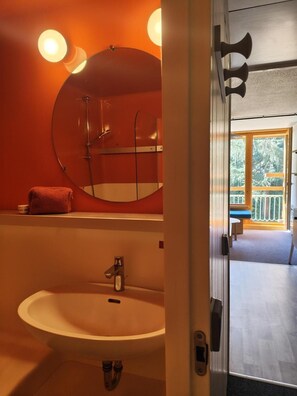 Ein Badezimmer mit rundem Spiegel, Waschbecken und Dusche. Ein Fenster bietet Blick ins Freie.