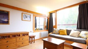 Una sala de estar acogedora con un sofá de madera, una mesa de centro y un mueble de madera con cajones. Hay dos ventanas con cortinas, un radiador y un cuadro enmarcado en la pared.