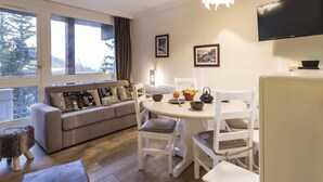 Una sala de estar acogedora con sofá, mesa de comedor y sillas. Hay ventanas con cortinas, un televisor de pantalla plana y cuadros en la pared.