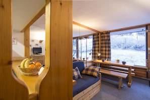A cozy cabin interior with a wooden bench, a sofa, a dining table, and a large window with plaid curtains.