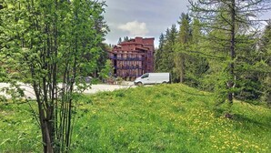 A building with a red facade and balconies, surrounded by lush greenery and a grassy area with yellow flowers.