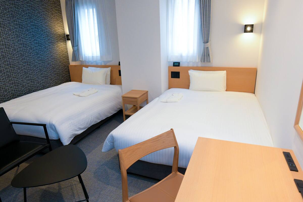 A hotel room with two single beds, a desk, a chair, and a small table.