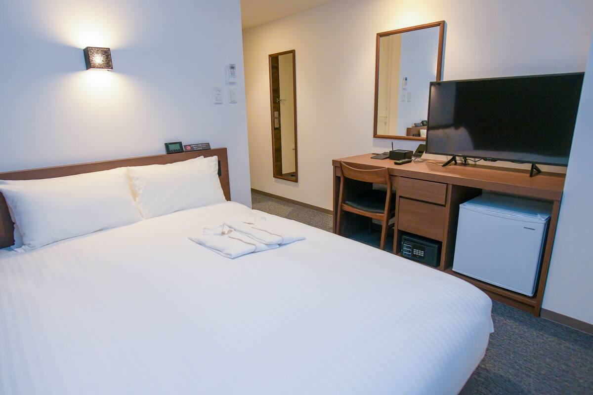 A hotel room with a bed, a desk with a television, a mirror, and a small refrigerator.