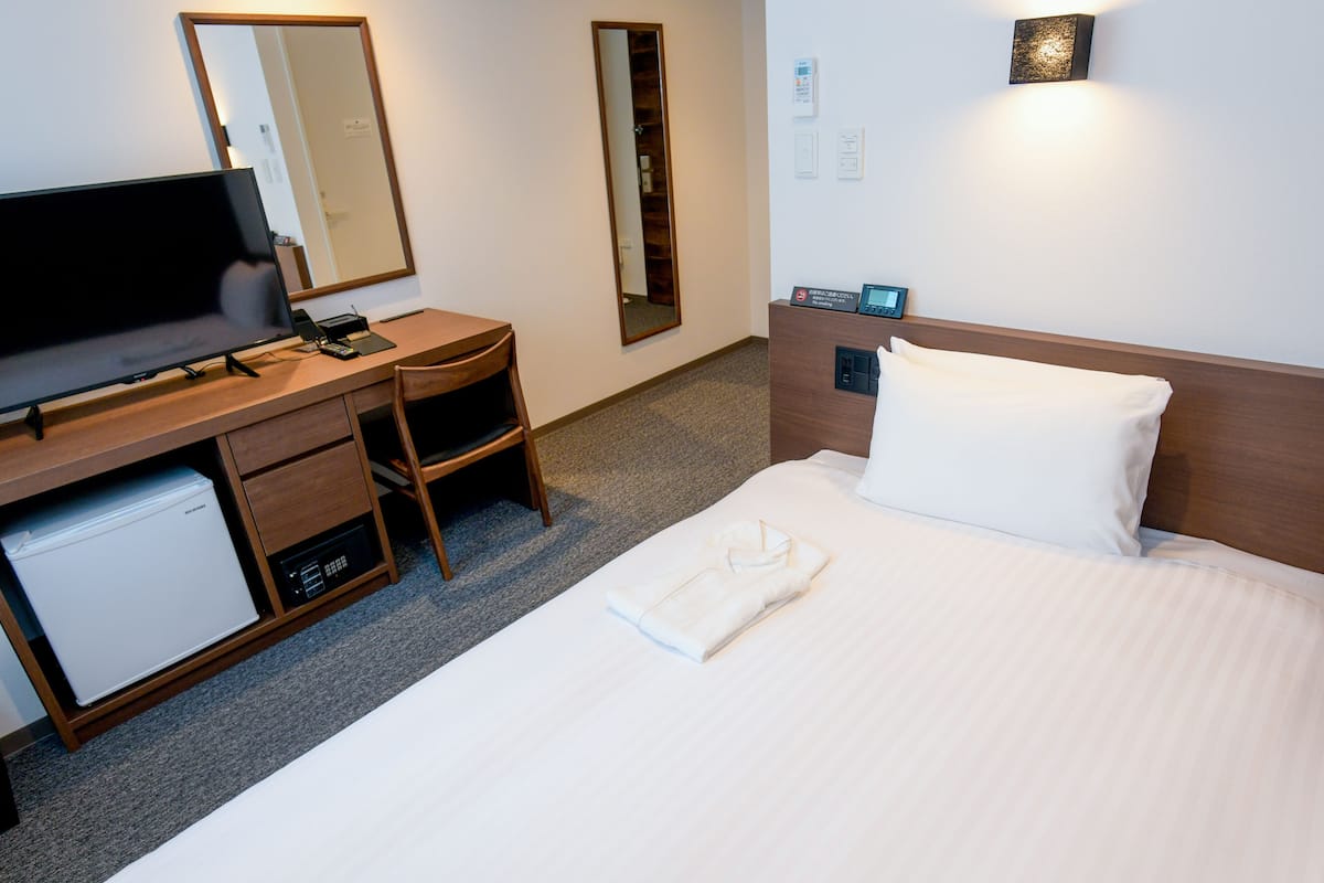A hotel room with a bed, a desk with a television, a refrigerator, a mirror, and a wall-mounted light.