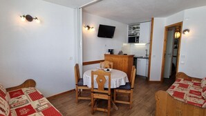 A cozy room with a wooden dining table and chairs, a television mounted on the wall, and a kitchen area visible through an open door. - 10m from ski slopes, view ski slopes, balcony, tv, 26m², La Plagne (Aime)