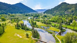 Un parcours de golf avec un pont enjambant une rivière, entouré d’une végétation luxuriante et de montagnes.