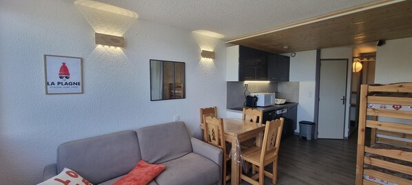 Un ambiente compatto con divano, tavolo da pranzo in legno e sedie. Si intravede in secondo piano una zona cucina con elettrodomestici moderni.