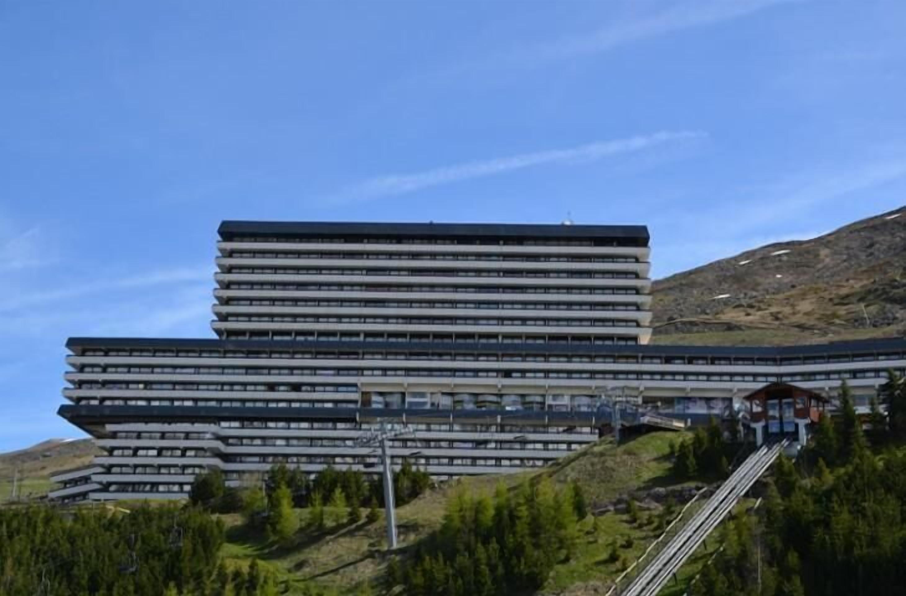 En moderne, flereetagers bygning med et unikt, trappetrin-lignende design, omgivet af et kuperet landskab og grøn vegetation.