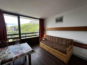 A room with a wooden sofa, a table with a newspaper, and a view of greenery and buildings through large windows.