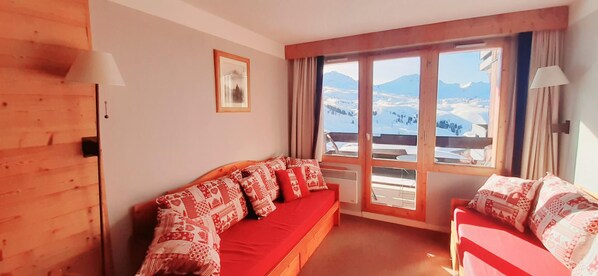 Um quarto aconchegante com um sofá de madeira e um sofá vermelho, janelas amplas com vista para as montanhas e um quadro na parede.