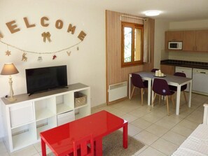 A compact living space with a TV on a white stand, a red table, a dining area with a white table and purple chairs, a kitchen with wooden cabinets and a microwave, and a 'WELCOME' sign on the wall. - 50m from shops, 1st floor, view valley, swimming pool, sauna, balcony, tv, ski locker, 34m² (Les Orres)