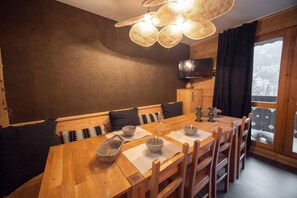 Un comedor con mesa de madera, sillas y un banco con cojines. Hay un televisor en la pared y una ventana con vistas.