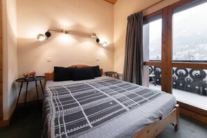 Un dormitorio con una cama grande, una mesita pequeña, una silla y una ventana que da a un paisaje nevado.