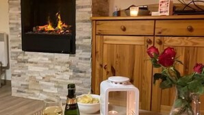 Una habitación con pared de piedra, chimenea, televisor, una mesa con una botella, un bol de patatas, una vela y un florero con rosas.