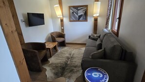 Ein gemütliches Wohnzimmer mit einer Couch, einem Sessel und einem kleinen Tisch. Ein Flachbildfernseher ist an der Wand befestigt und es gibt zwei Wandleuchten.
