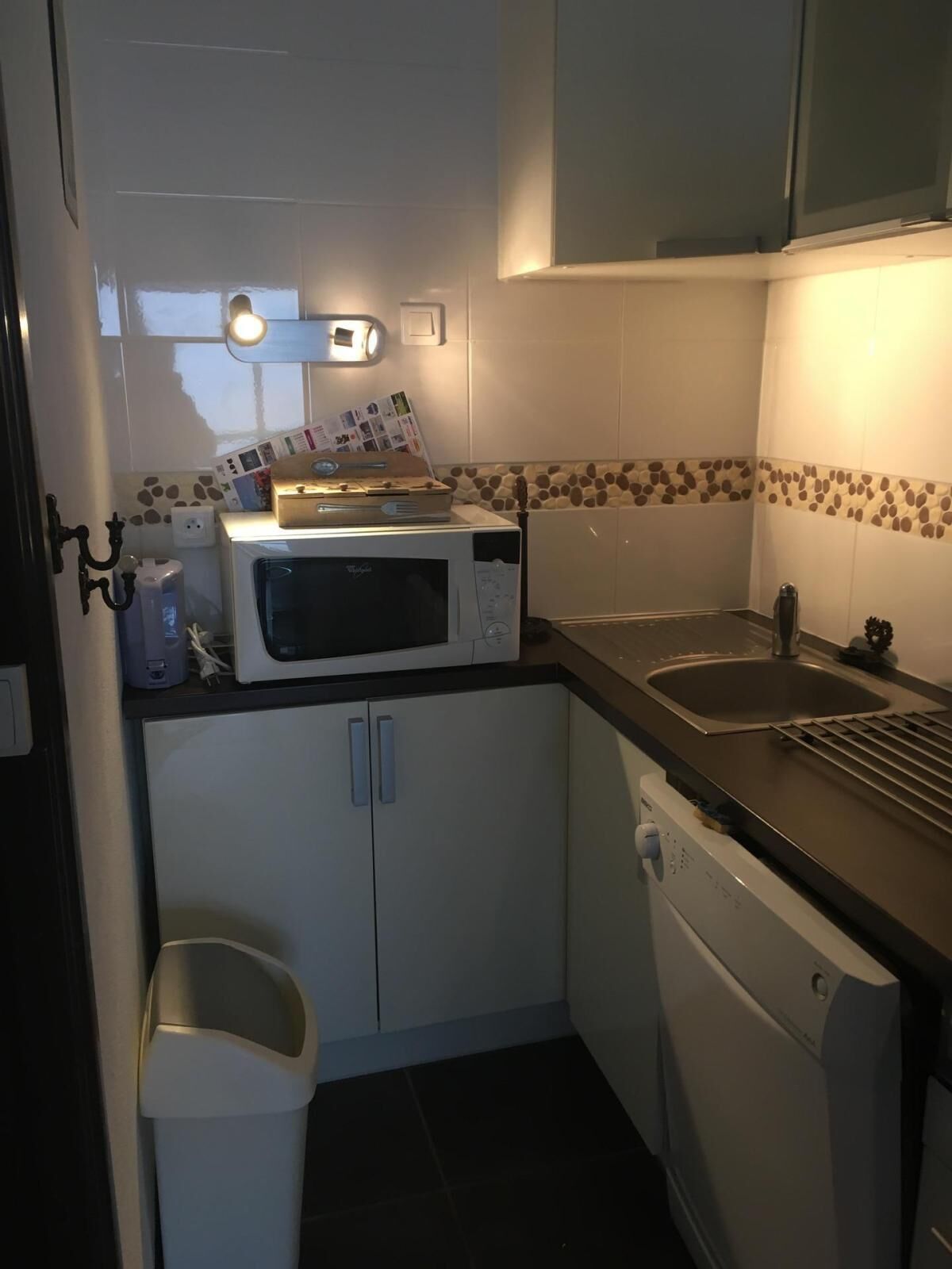 A compact kitchen with a microwave, a sink, and a dishwasher. There is a small trash bin and a cabinet with a tiled backsplash.