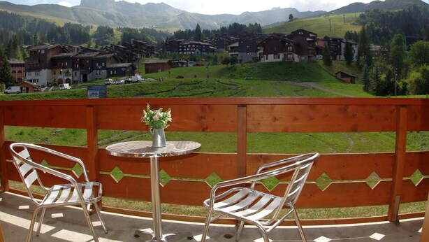 Ein Balkon mit zwei Metallstühlen und einem kleinen runden Tisch mit einem Blumenstrauß. Im Hintergrund eine Ansicht eines Dorfes mit Gebäuden und Bergen.