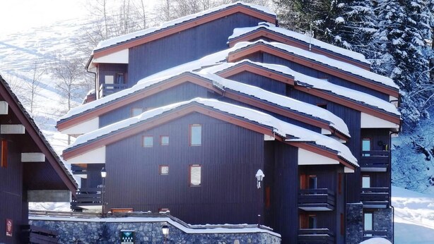 Un edificio de varios pisos con fachada oscura y varios balcones, rodeado de árboles nevados y montañas.