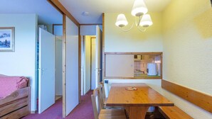 Een knus kamertje met een houten eettafel, bankjes, een kitchenette en een bed met een roze deken.