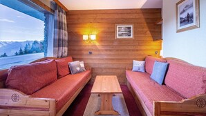 Een knusse kamer met twee houten banken, een houten tafel en uitzicht op bergen door een groot raam.