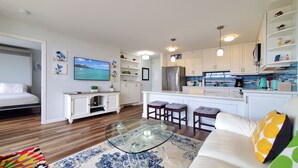 Una sala moderna con una mesa de centro de vidrio, un sofá y un televisor. Se observa un área de cocina con armarios blancos y un refrigerador.