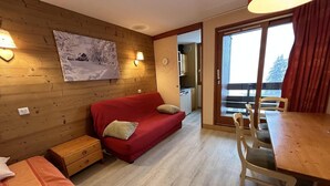 A cozy room with a red sofa, wooden walls, a dining table with chairs, and a view through a large window. - 75m from shops, 2nd floor, view valley, balcony, ski locker, 27m², Méribel-Mottaret (Méribel - Les Allues)