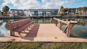Un muelle de madera con un banco, una masa de agua tranquila y un edificio con varios balcones al fondo.