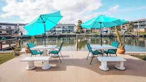 Un patio al aire libre con mesas y sillas, sombrillas grandes y un cuerpo de agua al fondo.
