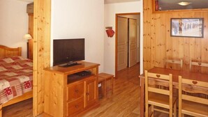 A wooden-floored room with a bed, a TV on a wooden stand, a dining table with chairs, and a wooden wall with a framed picture.
