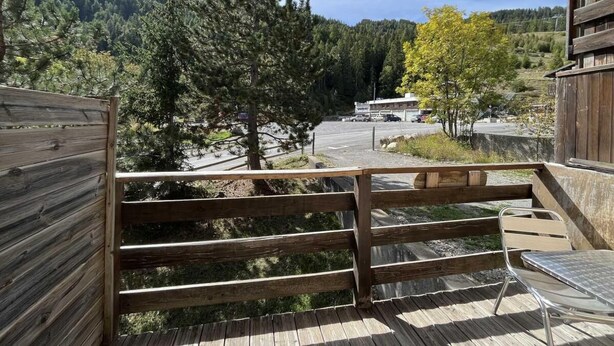 A wooden deck with a metal chair, overlooking a parking area and a forested hillside.