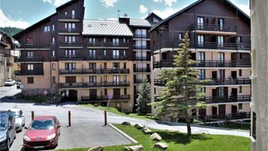 A multi-story building with balconies, a parking area with cars, and a grassy area with rocks. - 100m from ski slopes, 1st floor, balcony, tv, ski locker, 28m², Risoul (Risoul)