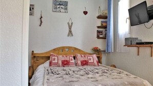 A bedroom with a wooden bed, patterned pillows, a wall-mounted TV, and a shelf with books and decorative items. - 100m from ski slopes, 1st floor, balcony, tv, ski locker, 28m², Risoul (Risoul)