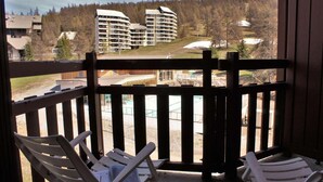 Un balcon doté de balustrades en bois et de deux fauteuils de jardin blancs. En arrière-plan, on aperçoit des immeubles à plusieurs étages et une vue sur une zone boisée.