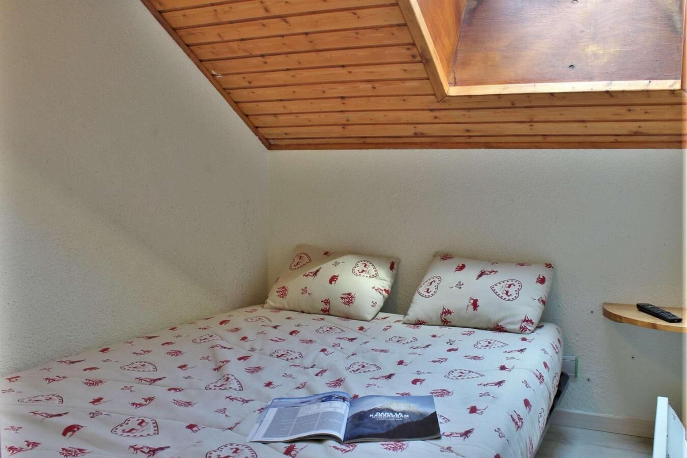 Quarto com teto inclinado, cama com roupa de cama estampada, duas almofadas, mesa de cabeceira com controle remoto e um livro na cama.