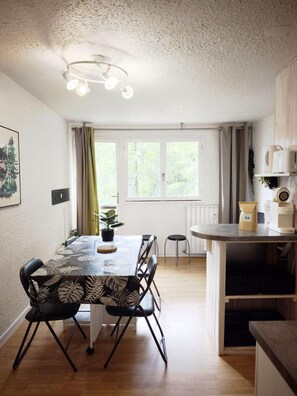 Zona pranzo con tavolo, sedie, lampadario, finestra con tende e piano cucina con elettrodomestici.