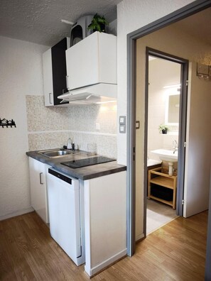 A compact kitchen with a dishwasher, a sink, and a microwave. There is a small window above the sink, and a door leading to a bathroom.